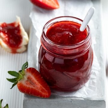 Open jar of strawberry jam, with spoon sticking out of it, sitting on a grey plate with a strawberry on the edge.