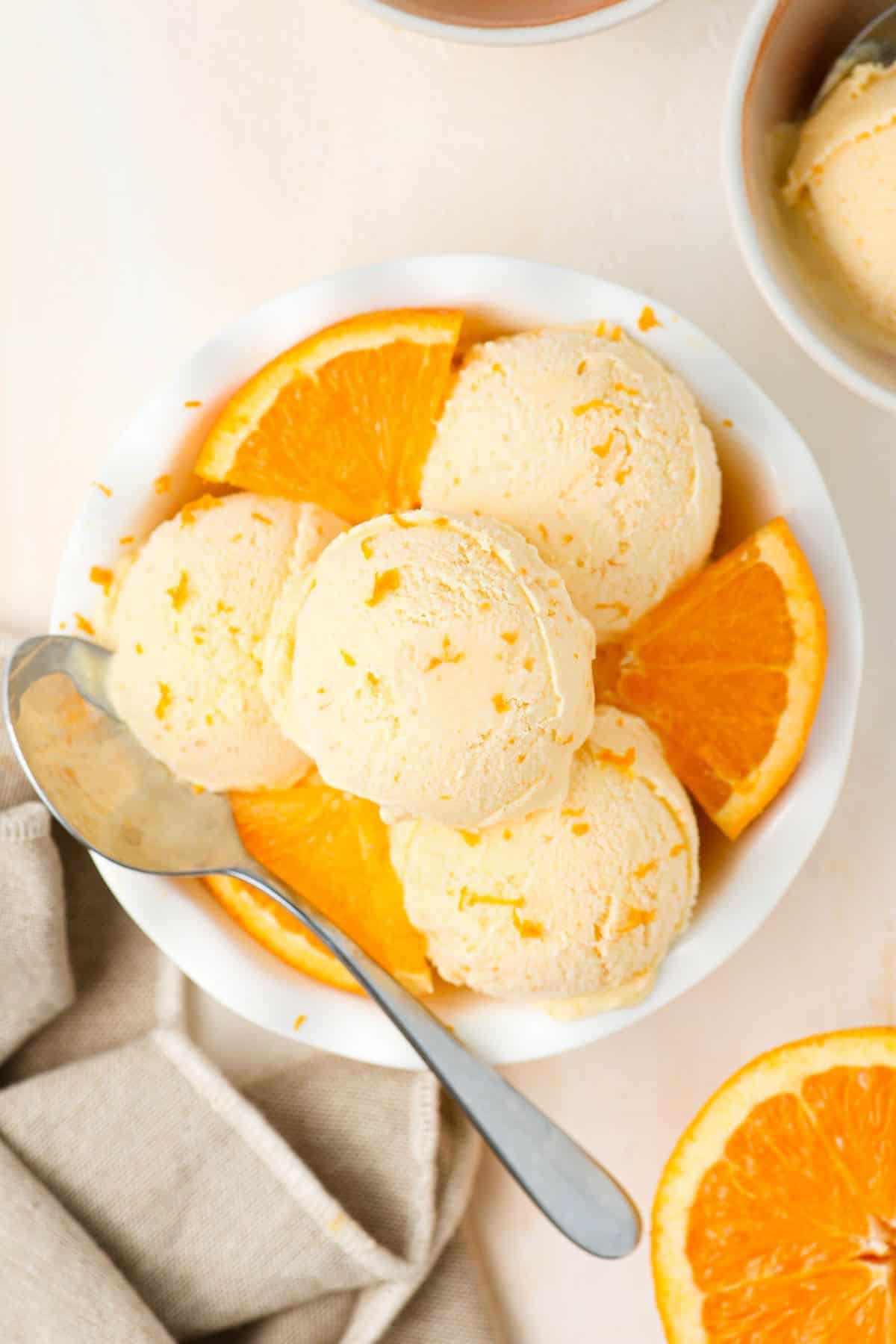 Round white bowl of ice cream with four scoops and some orange pieces, and a spoon on the edge.