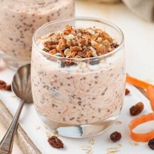 Glass of overnight oats garnished with walnuts and grated carrot, sitting on board with a spoon on the edge.