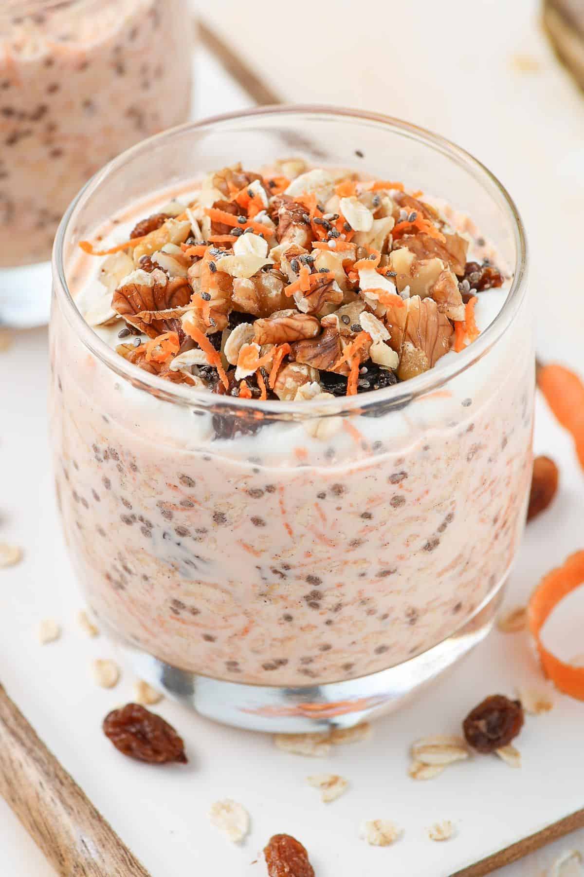Glass of overnight oats garnished with walnuts and grated carrot, sitting on a board.