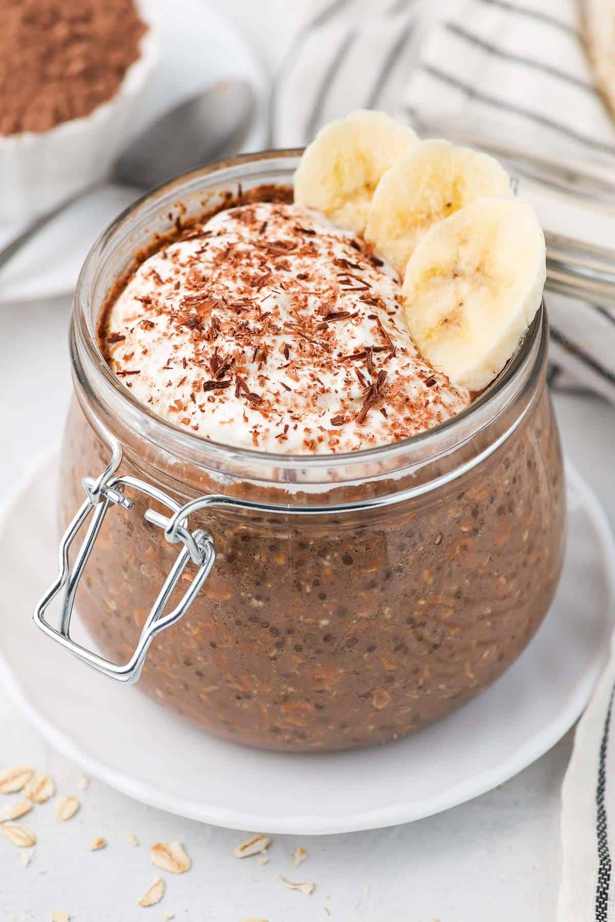 Jar of oats, topped with yoghurt, slices of banana and shaved chocolate.