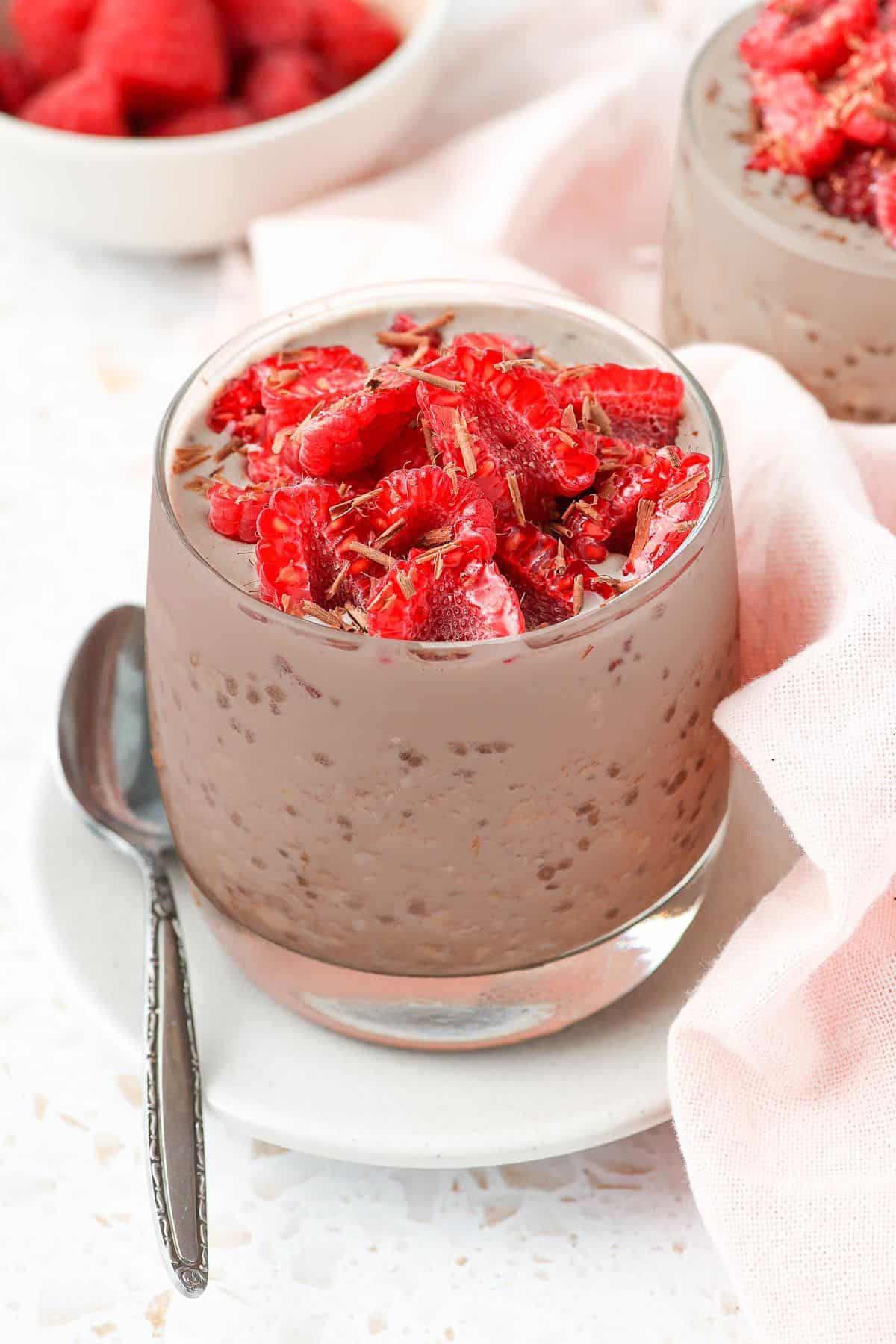 Jar of overnight oats, garnished with raspberries and chocolate shavings sitting on a small plate with a spoon on the edge.