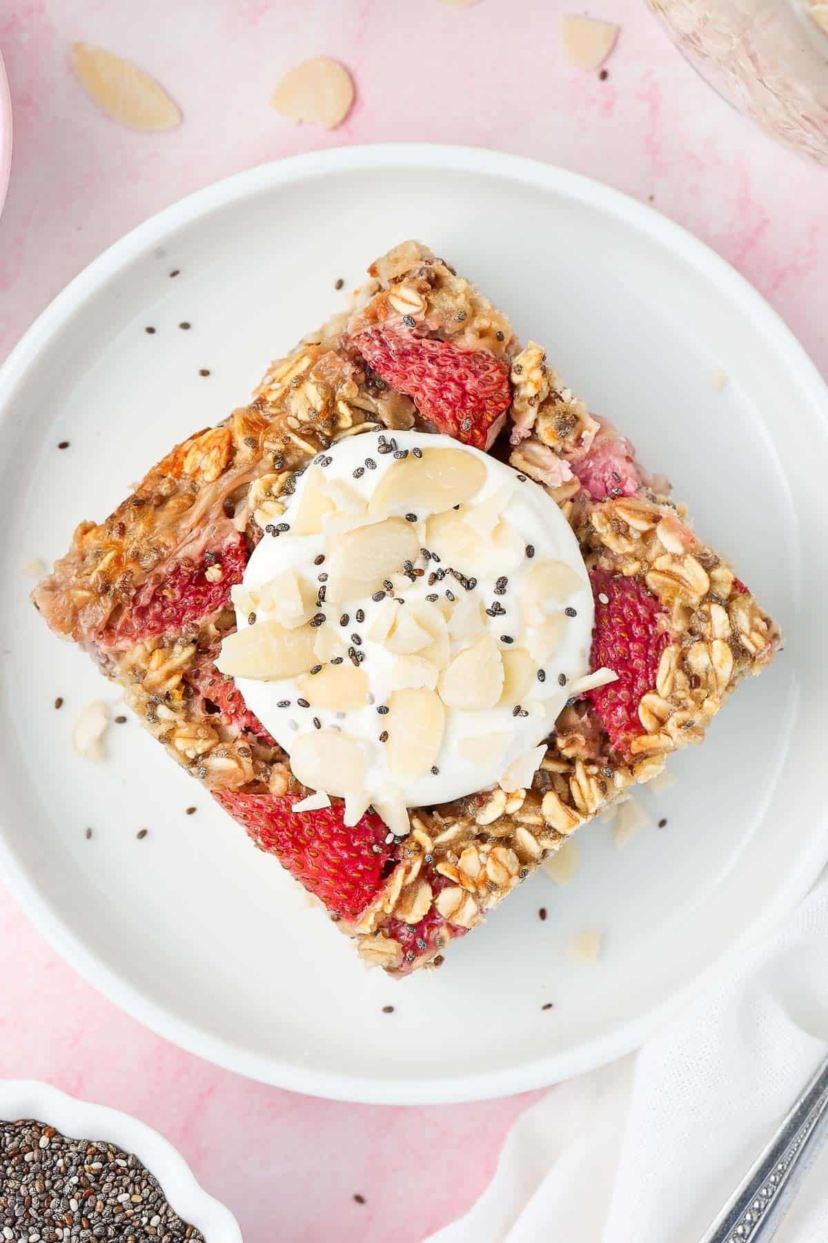 Round white plate with a piece of baked strawberry oats, topped with some yoghurt and flaked almonds.