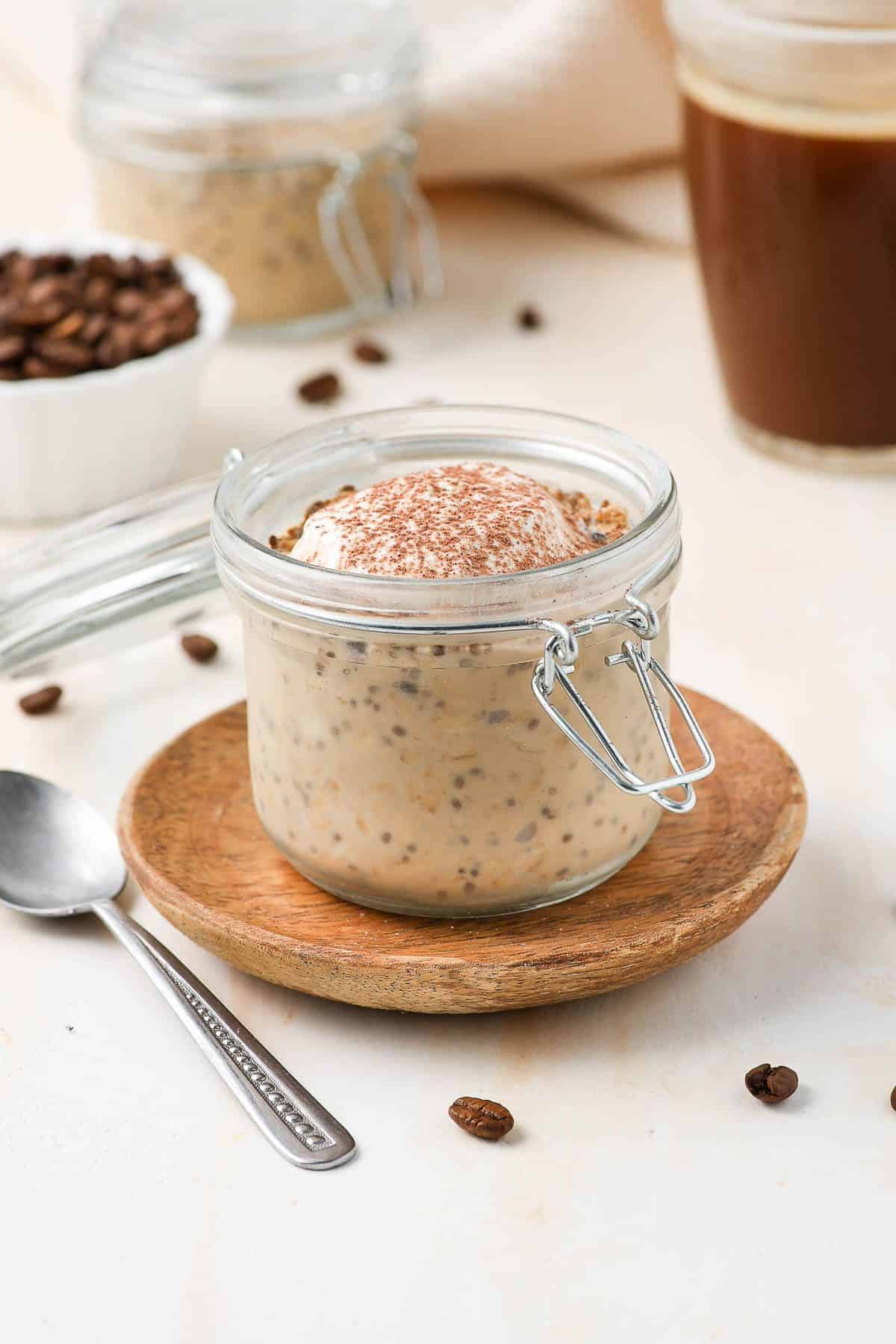 Jar of overnight oats, garnished with yoghurt and cocoa powder, sitting on a wooden saucer with a spoon on edge.