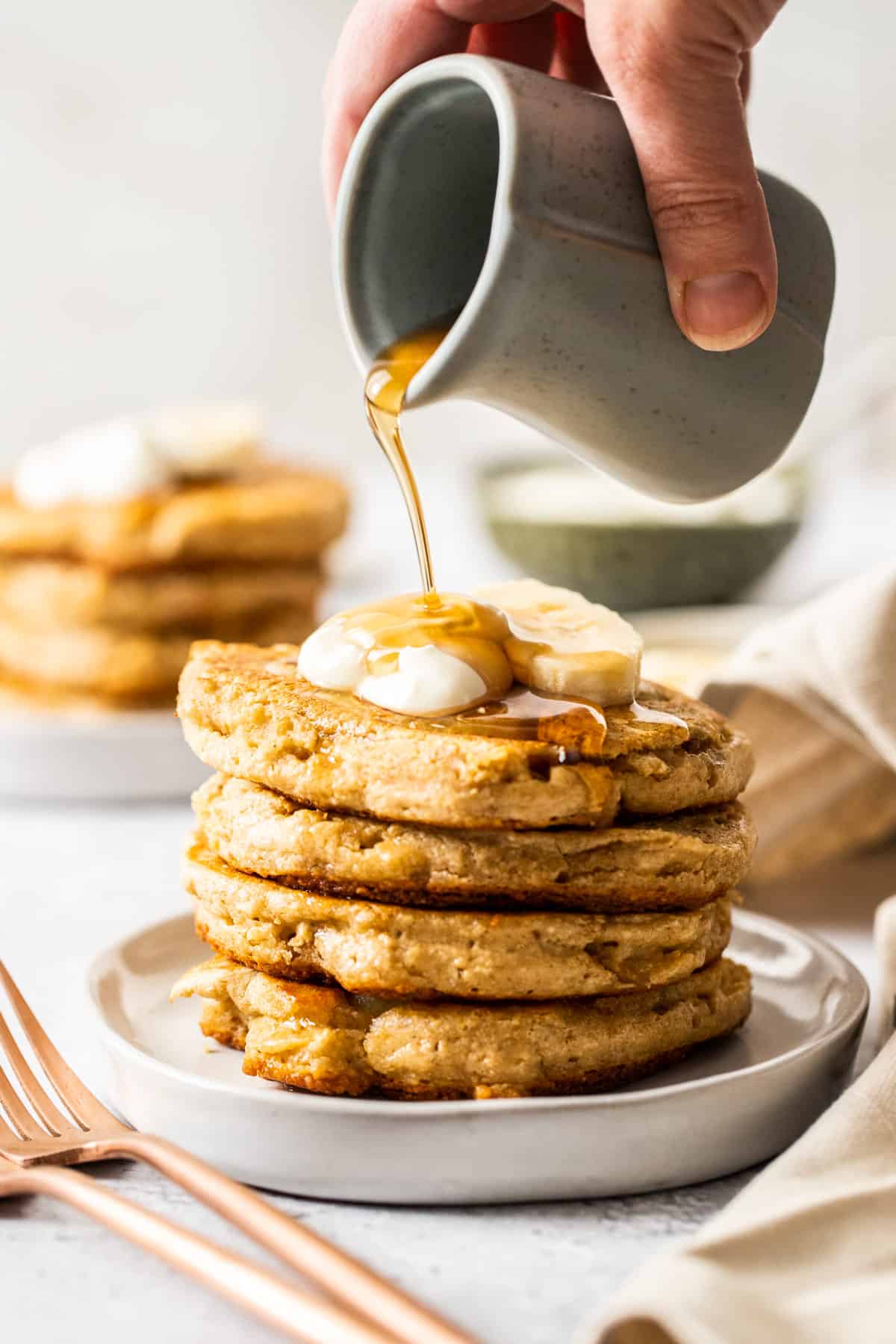 Stack of pancakes on a plate, topped with yoghurt, sliced banana and maple syrup being poured out of a jug.