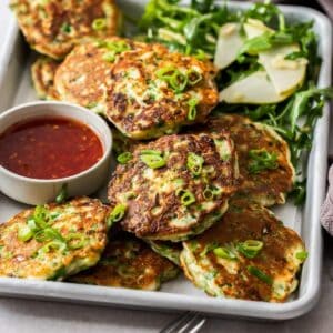 Sheet pan with cooked fritters, a small dish of chilli sauce and a rocket salad.