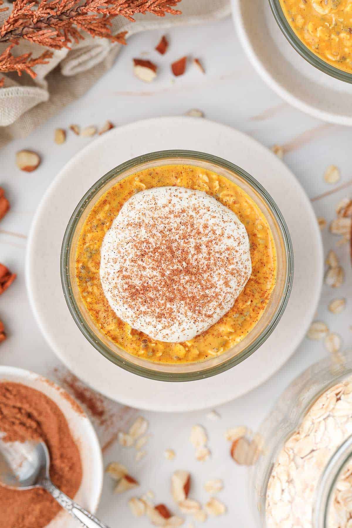 Overhead shot of glass jar of overnight oats topped with yoghurt and pumpkin spice, sitting on a white saucer.