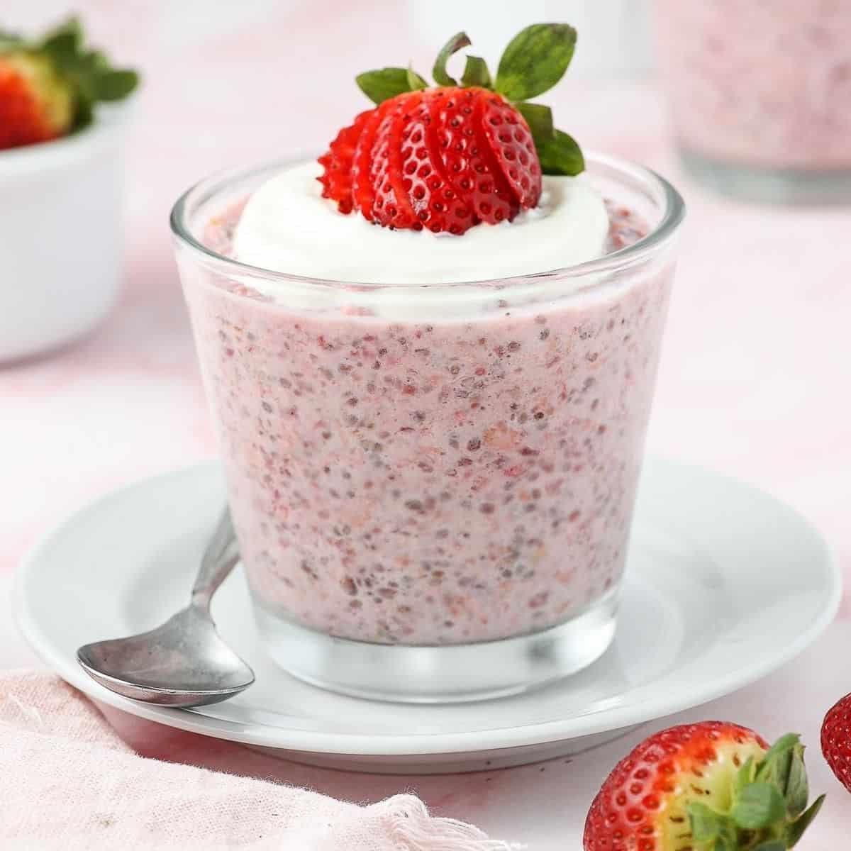 Glass jar of strawberry overnight oats, topped with yoghurt and a strawberry, sitting on a white saucer with a spoon on edge.