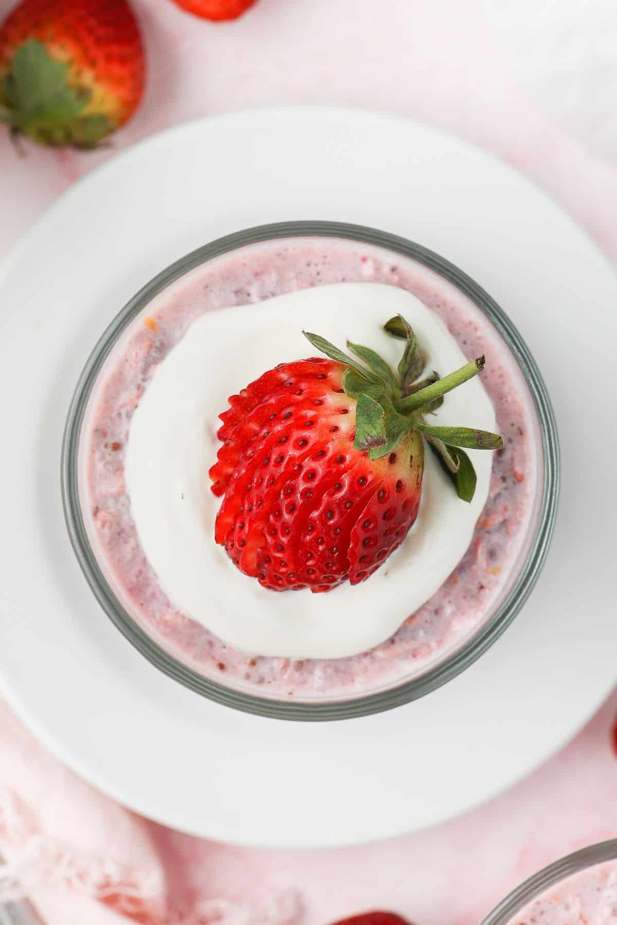 Glass jar of strawberry overnight oats, topped with yoghurt and a strawberry, sitting on a white saucer.