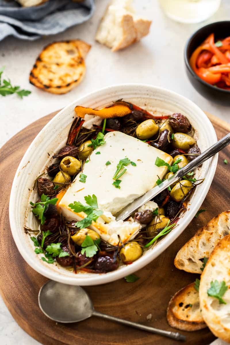 Oval dish of baked feta cheese with olives sitting on a wooden board, with a spoon and knife.