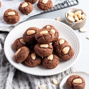 Round white plate with chocolate almond cookies, sitting on a grey cloth.