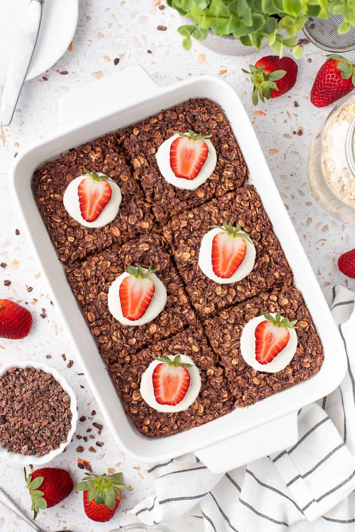 White rectangular dish of baked chocolate oatmeal, cut into six pieces, with a spoon of yoghurt and strawberry half on each piece.