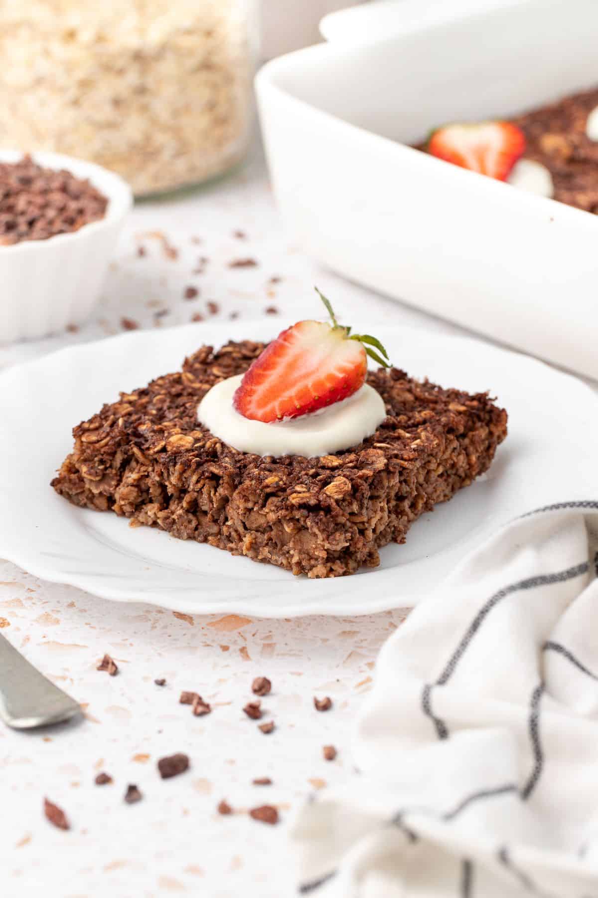 Small round white plate, with piece of oatmeal, with a spoon of yoghurt and half strawberry on top, and fork on edge of plate.