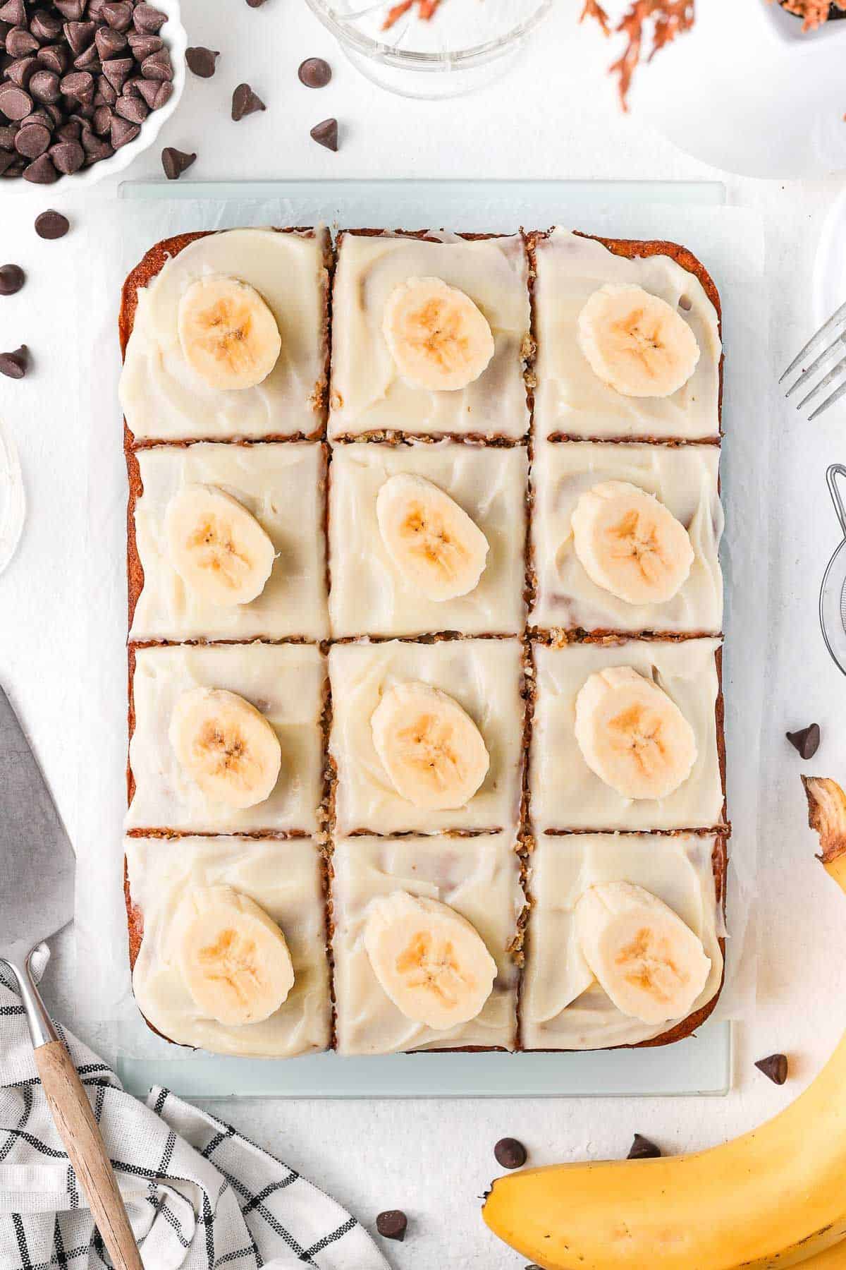 Banana Slice with cream cheese frosting, cut into 12 squares and topped with a slice of fresh banana, sitting on a glass board.