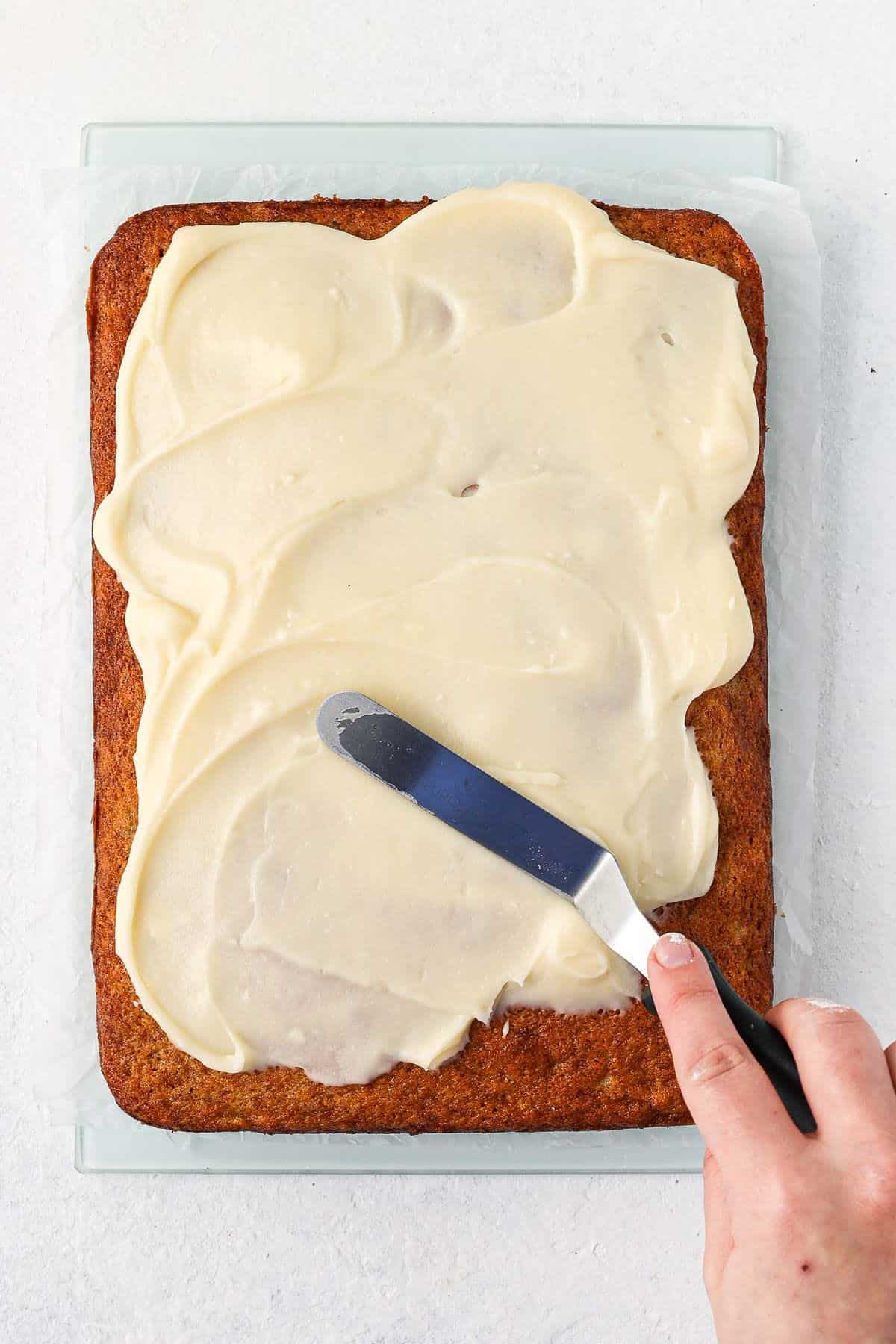 Banana Slice being iced with cream cheese frosting, showing a hand with a palette knife smoothing frosting.