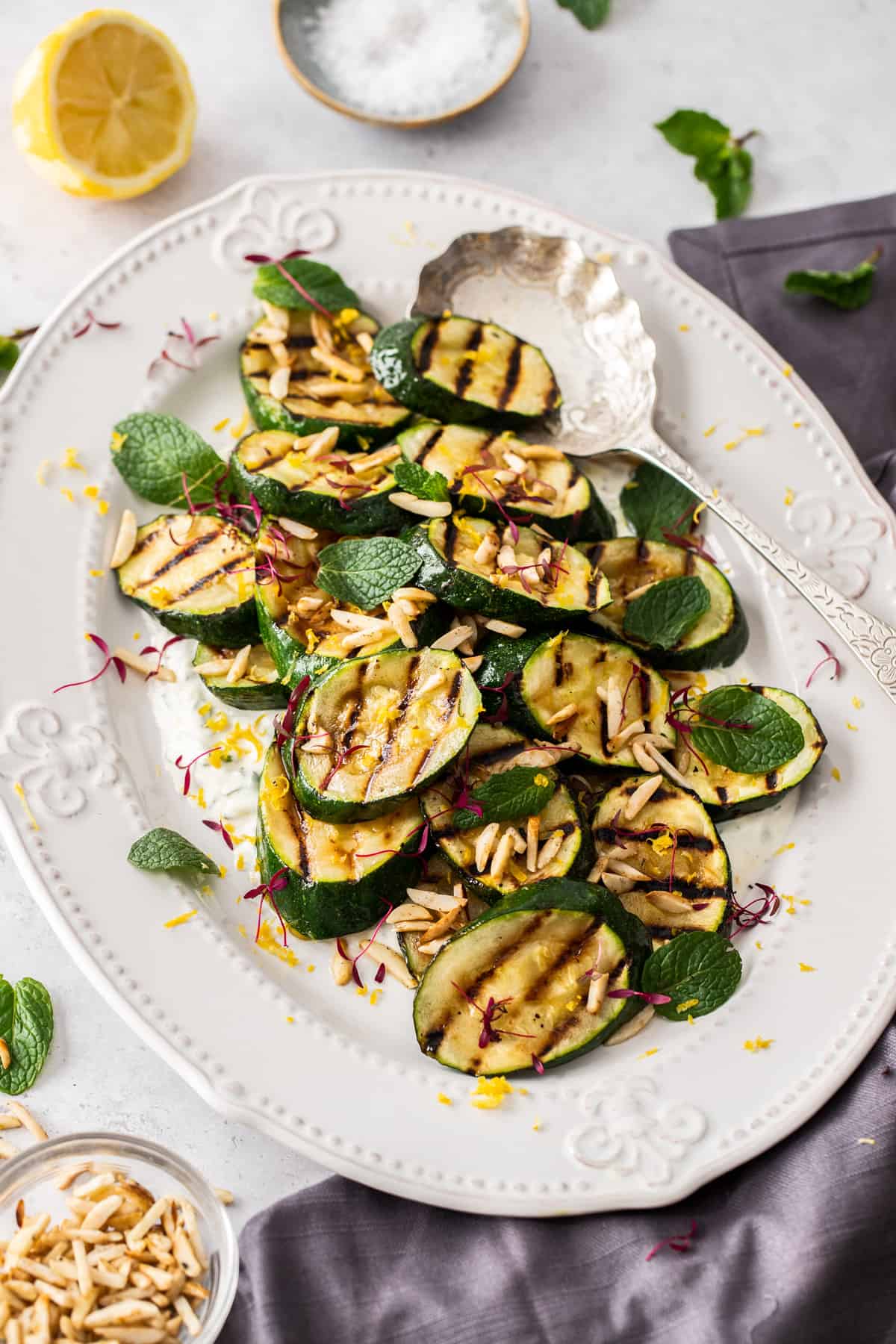 Oval white platter with grilled zucchini, garnished with slivered almonds, with a silver serving spoon on edge of the plate.
