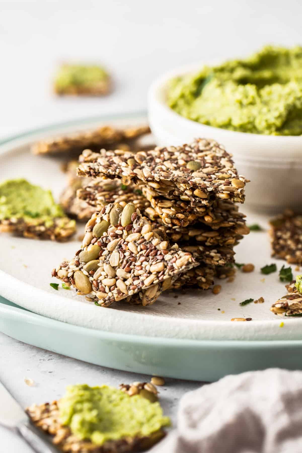 Stack of mixed seed crackers sitting on a white plate, with a dish of pea dip in the background.