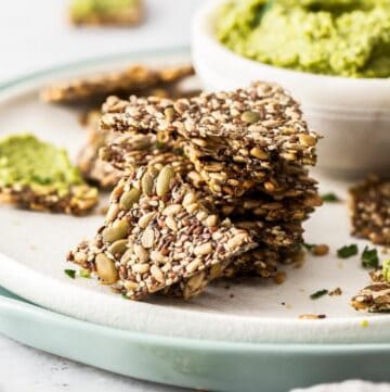Stack of mixed seed crackers sitting on a white plate, with a dish of pea dip in the background.