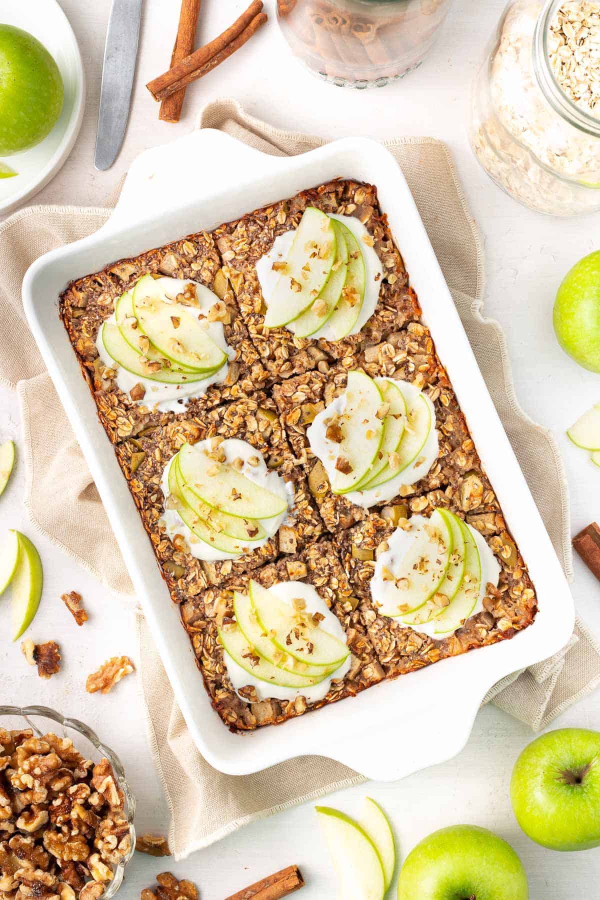 White rectangular dish of baked apple oatmeal, cut into six pieces and topped with yoghurt and apple slices.
