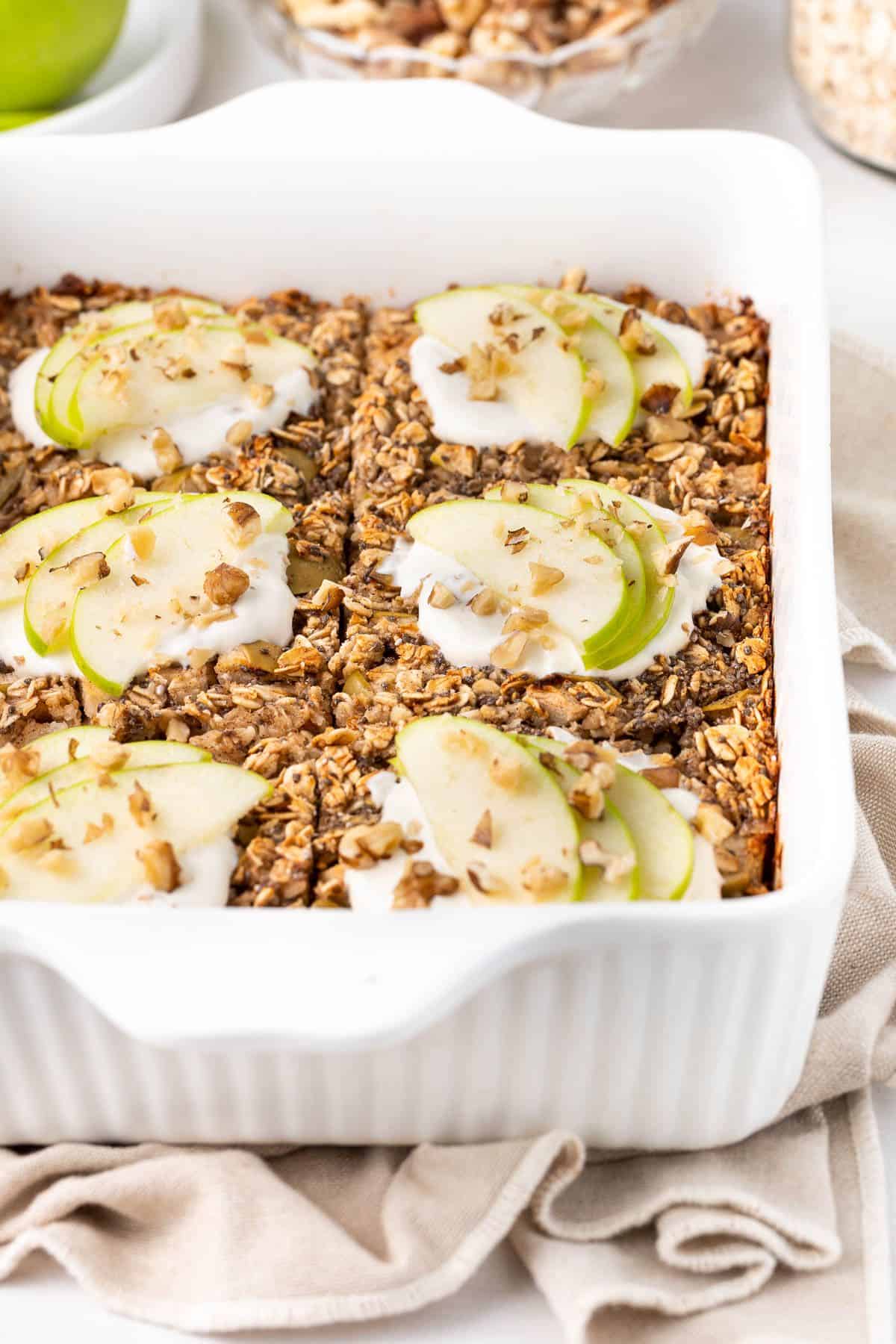 White rectangular dish of baked apple oatmeal, cut into six pieces and topped with yoghurt and apple slices.