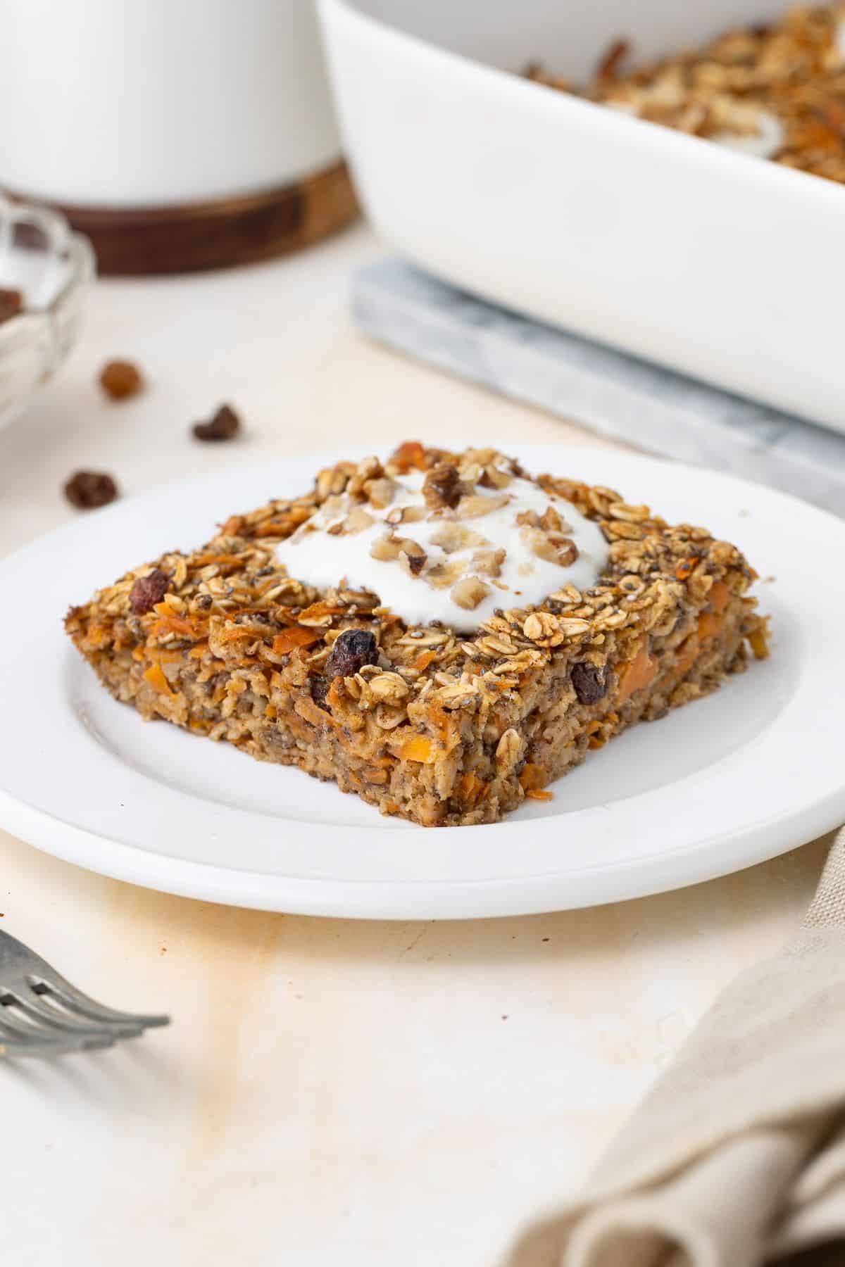 Piece of baked carrot oatmeal, sitting on a round white plate, topped with yoghurt and chopped walnuts.