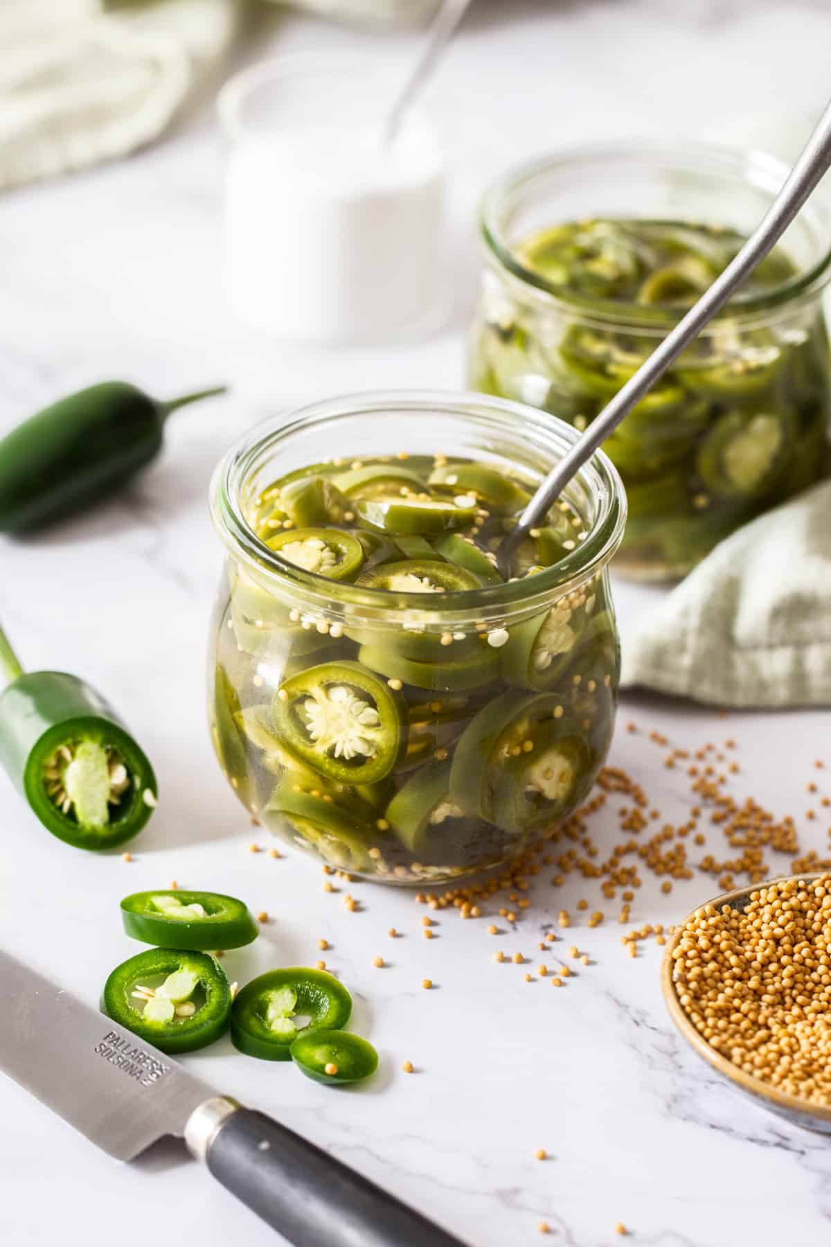 Jar of pickled jalapeños with a spoon sitting in jar, with some fresh jalapeños around the edge.