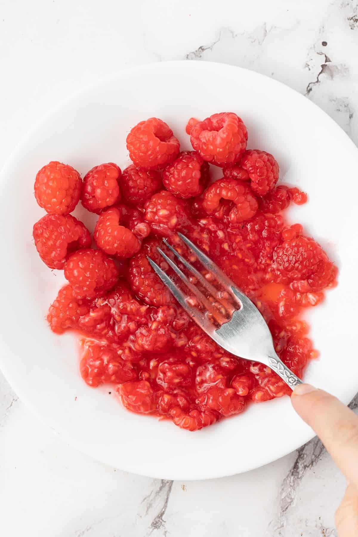 Round white dish with raspberries being mashed with a fork.