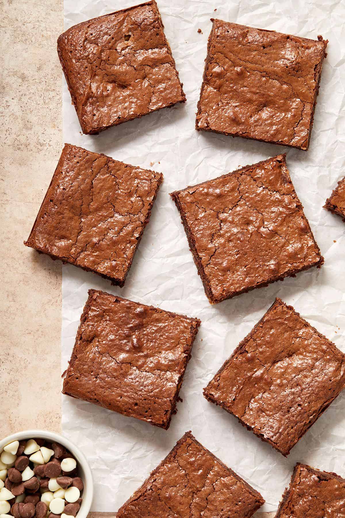 Overhead shot of squares of brownies on parchment paper, with a dish of chocolate chips in the bottom left corner.