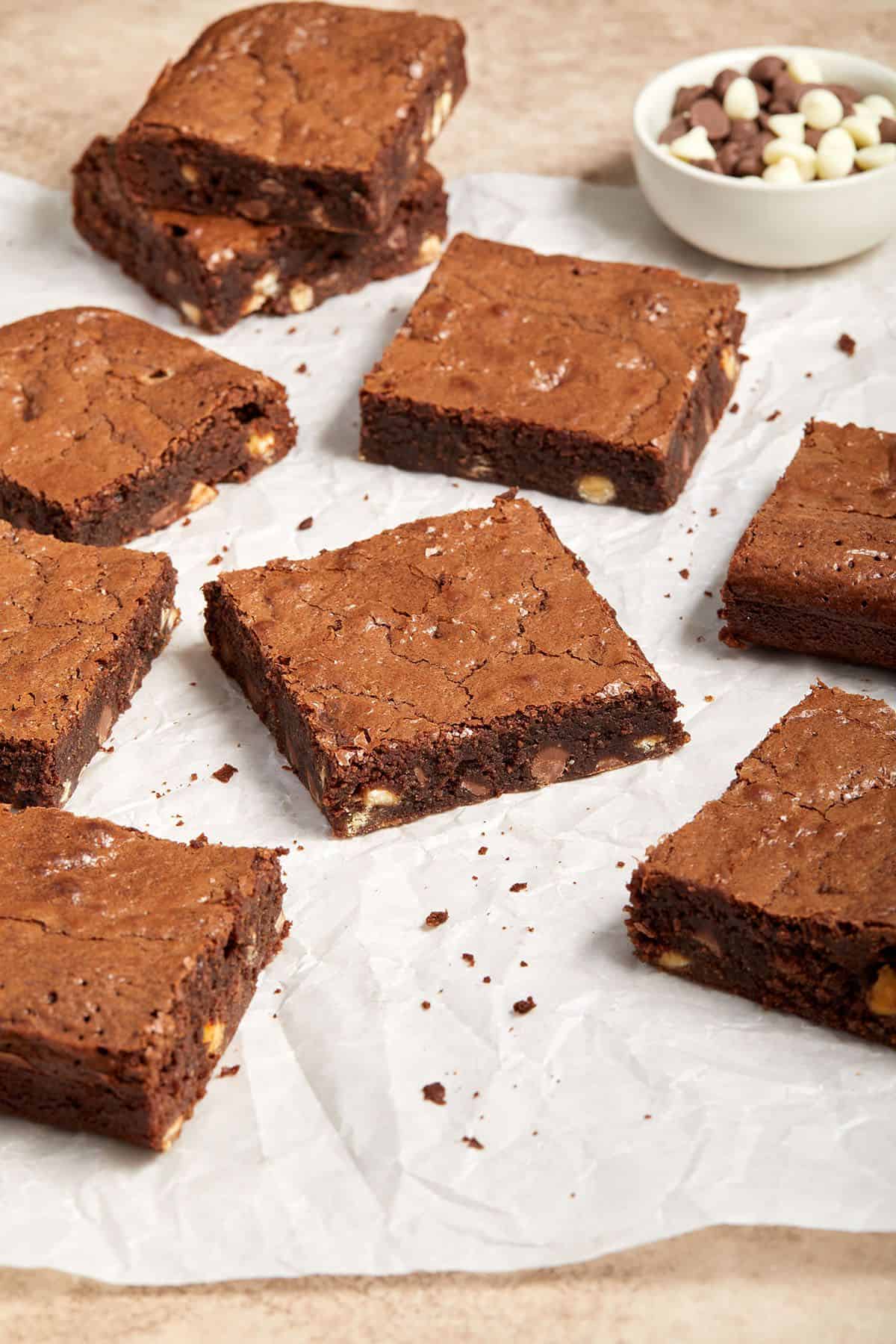 Cut squares of brownies on parchment paper, with a small dish of chocolate chips in the top right corner.
