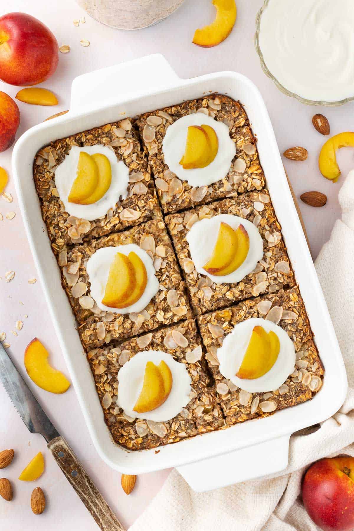 White rectangular dish of baked peach oatmeal, cut into six pieces and topped with yoghurt and peach slices.