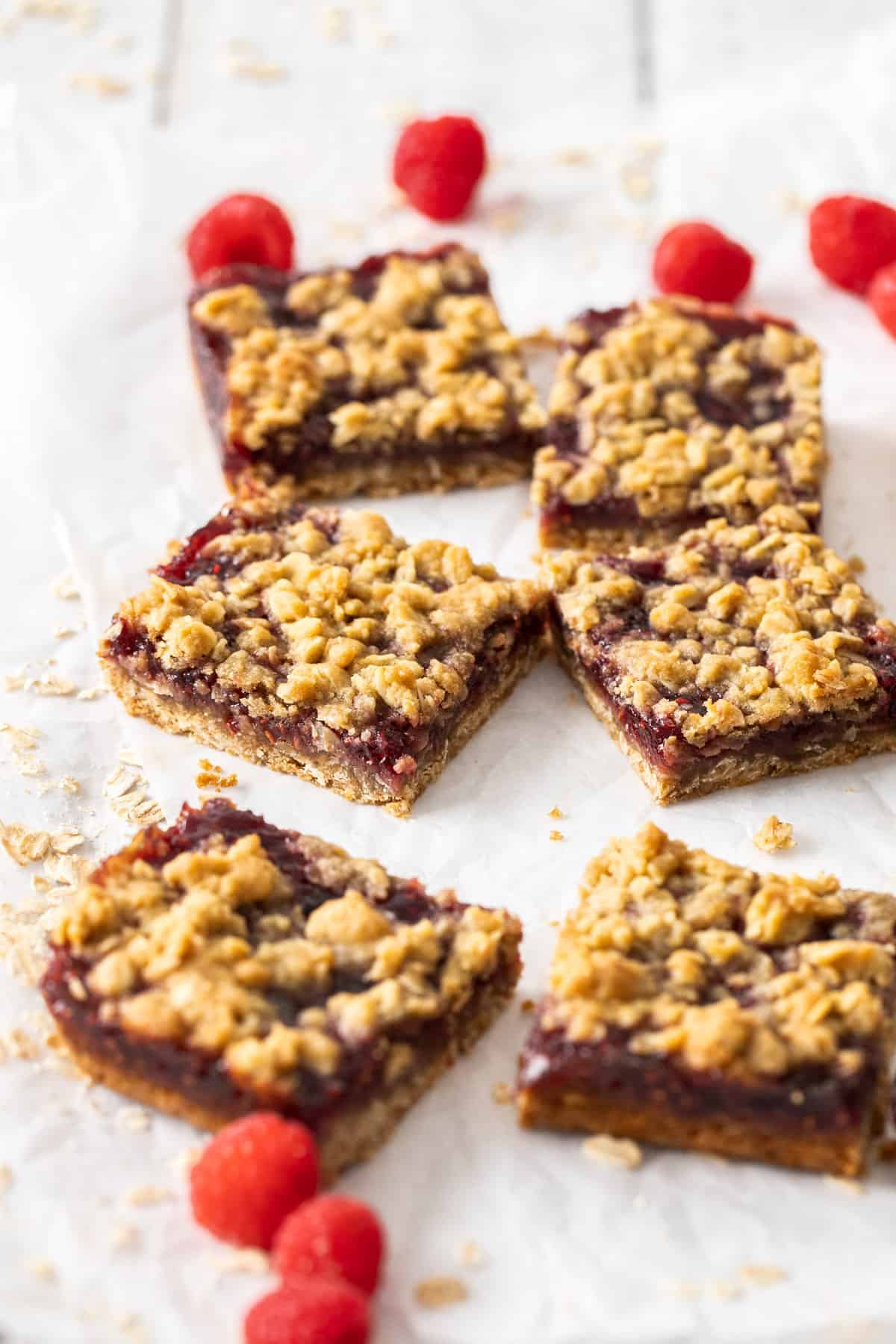 Cut squares of jam bars, sitting on parchment paper with a few raspberries scattered around the edge.