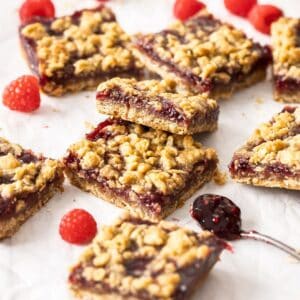 Cut squares of jam bars, sitting on parchment paper with a few raspberries scattered around the edge.