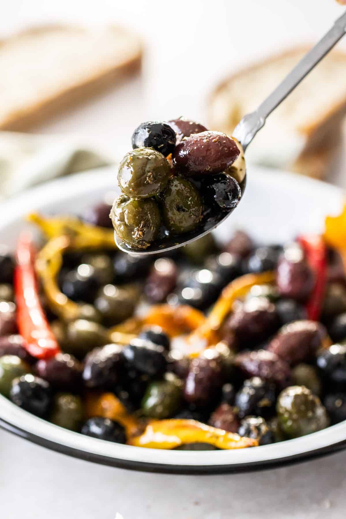 Spoon holding up a scoop of roasted olives, with the dish of olives in the background.
