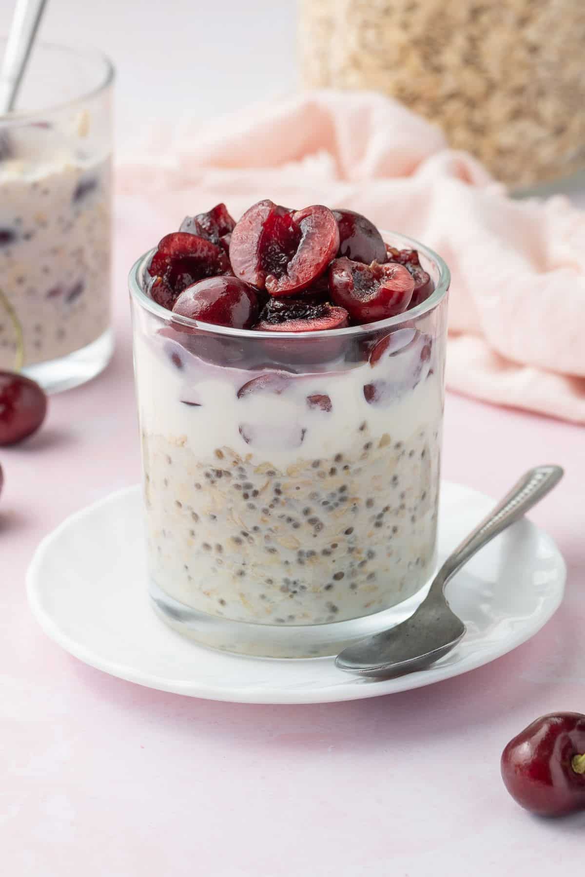 Glass jar of vanilla overnight oats, topped with halved cherries, sitting on a small white saucer with a teaspoon on the edge.