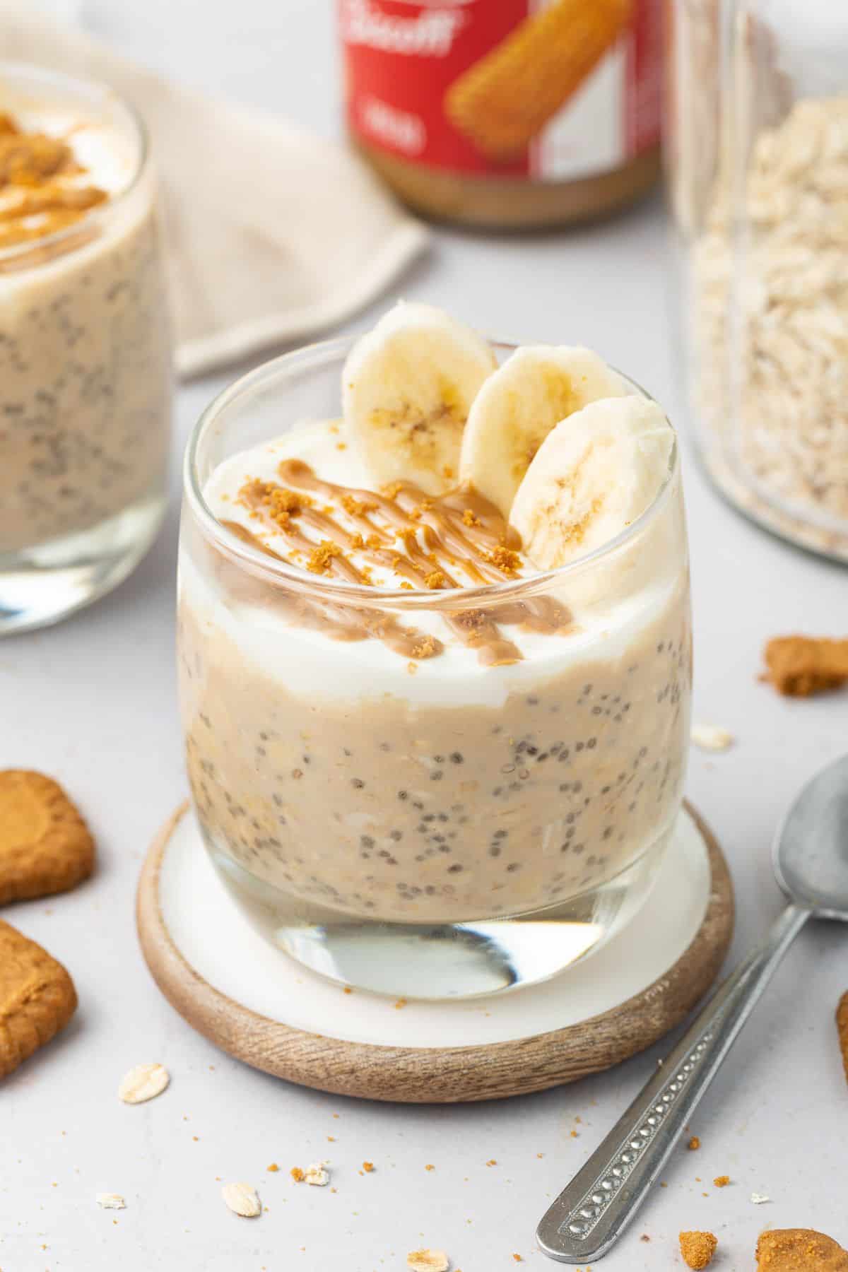 Glass jar of overnight oats topped with yoghurt, slices of banana and a drizzle of Biscoff spread, sitting on a round coaster.