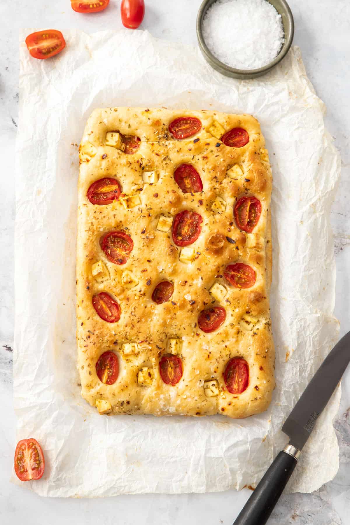 Overhead shot of spelt focaccia, studded with feta and tomatoes, sitting on a piece of baking paper.