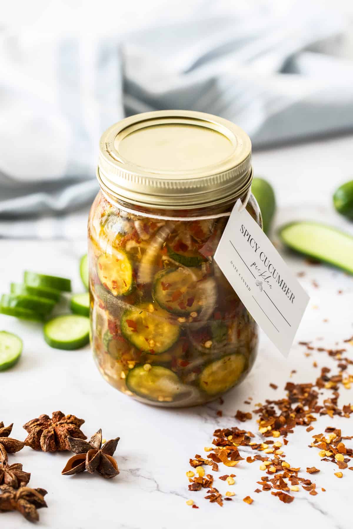 Jar of Spicy Cucumber Pickles, sealed with gold lid, and a label around the top, surrounded by spices and cucumber slices.