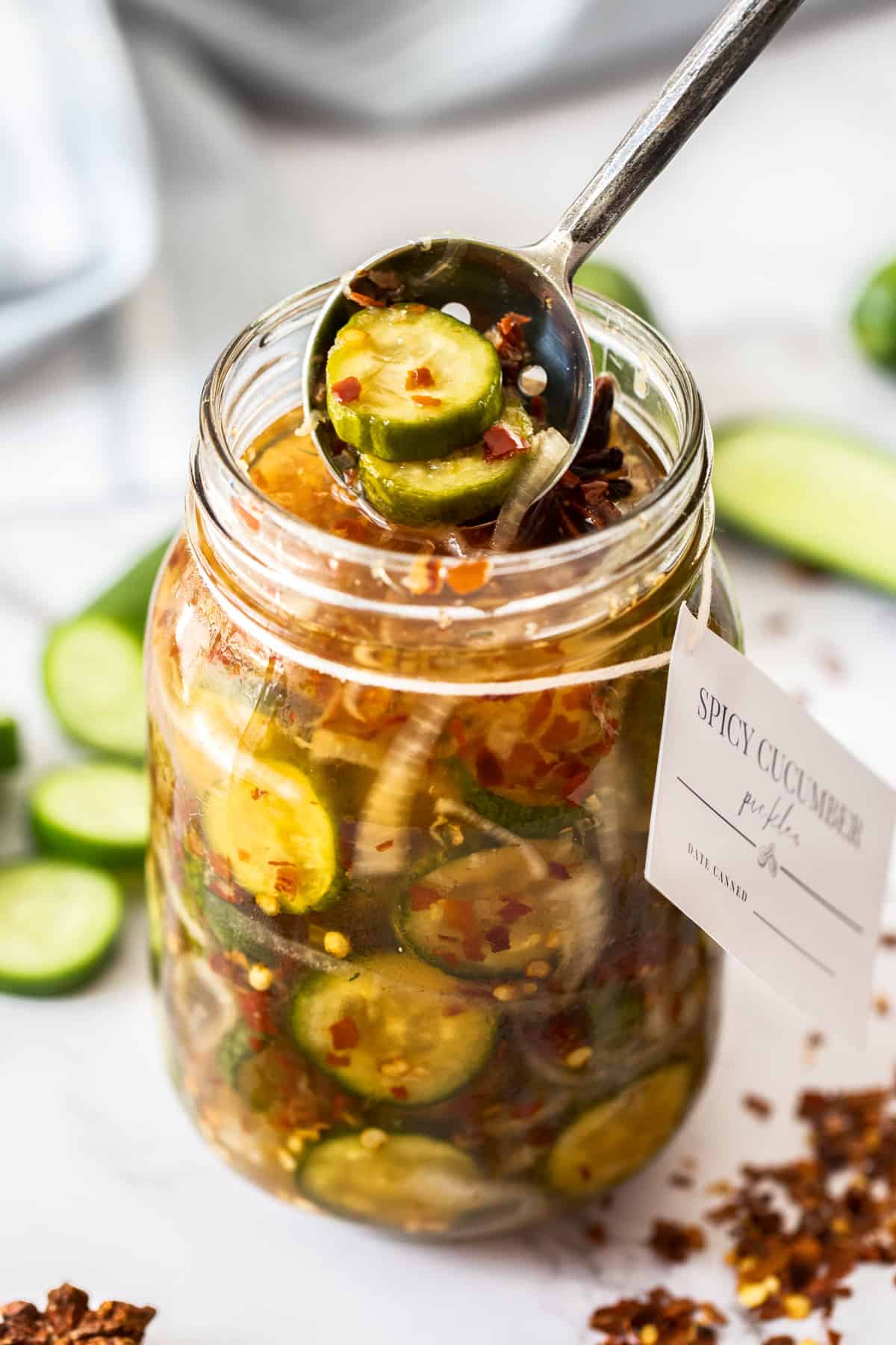 Jar of Spicy Cucumber Pickles, with a spoon reaching in to lift some pickles out of the jar.