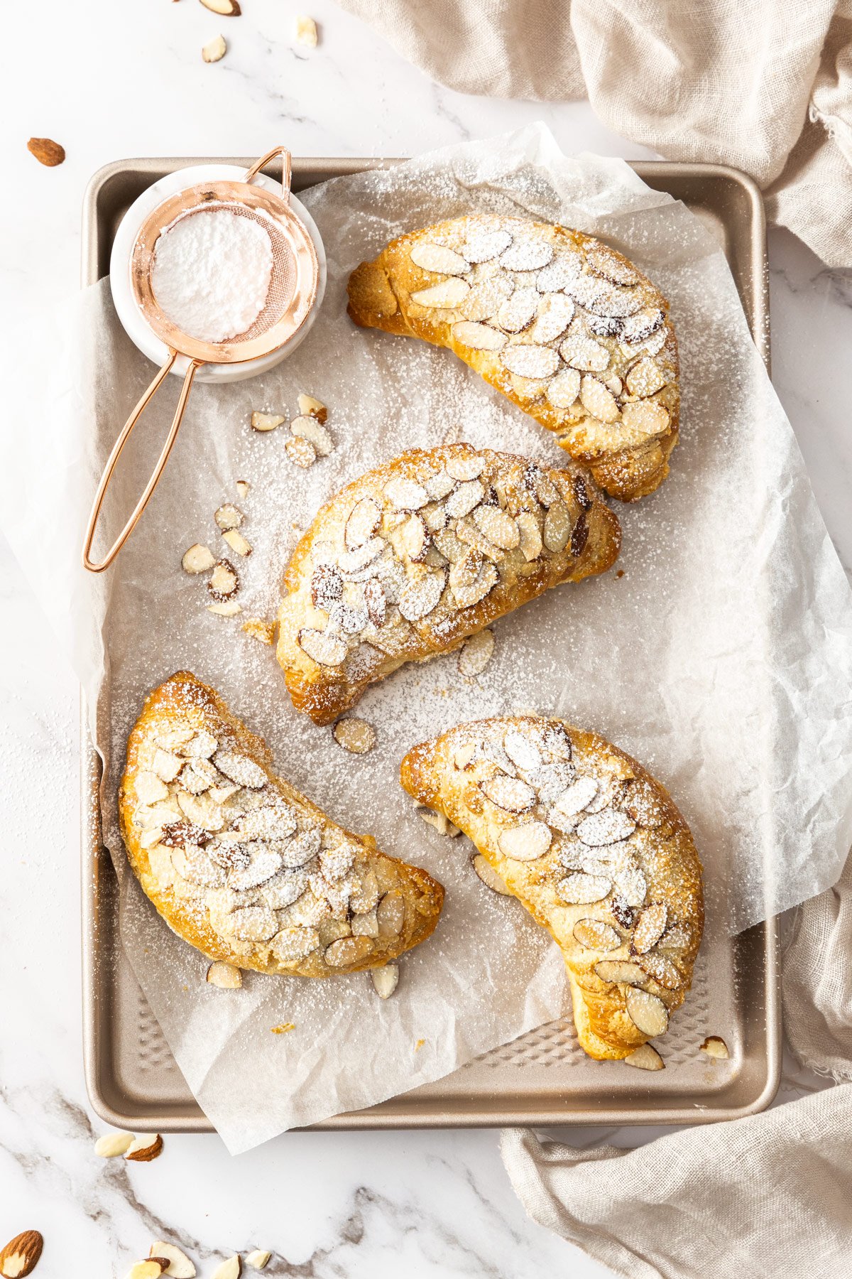 Four Almond Croissants, dusted with powdered sugar, sitting on some baking paper on a gold baking tray.