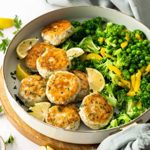 Round white dish of chicken patties, with some broccoli, peas and peppers served on the side.