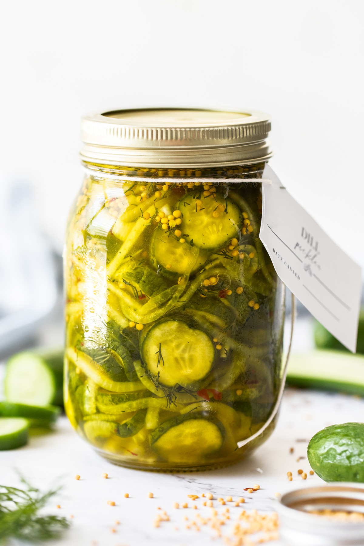 Jar of Dill Cucumber Pickles, sealed with gold lid, and a label around the top, surrounded by spices and cucumber slices.