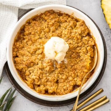 Round white dish of Pineapple Crisp, with a scoop of ice cream in the centre, and a gold spoon resting in the dish.