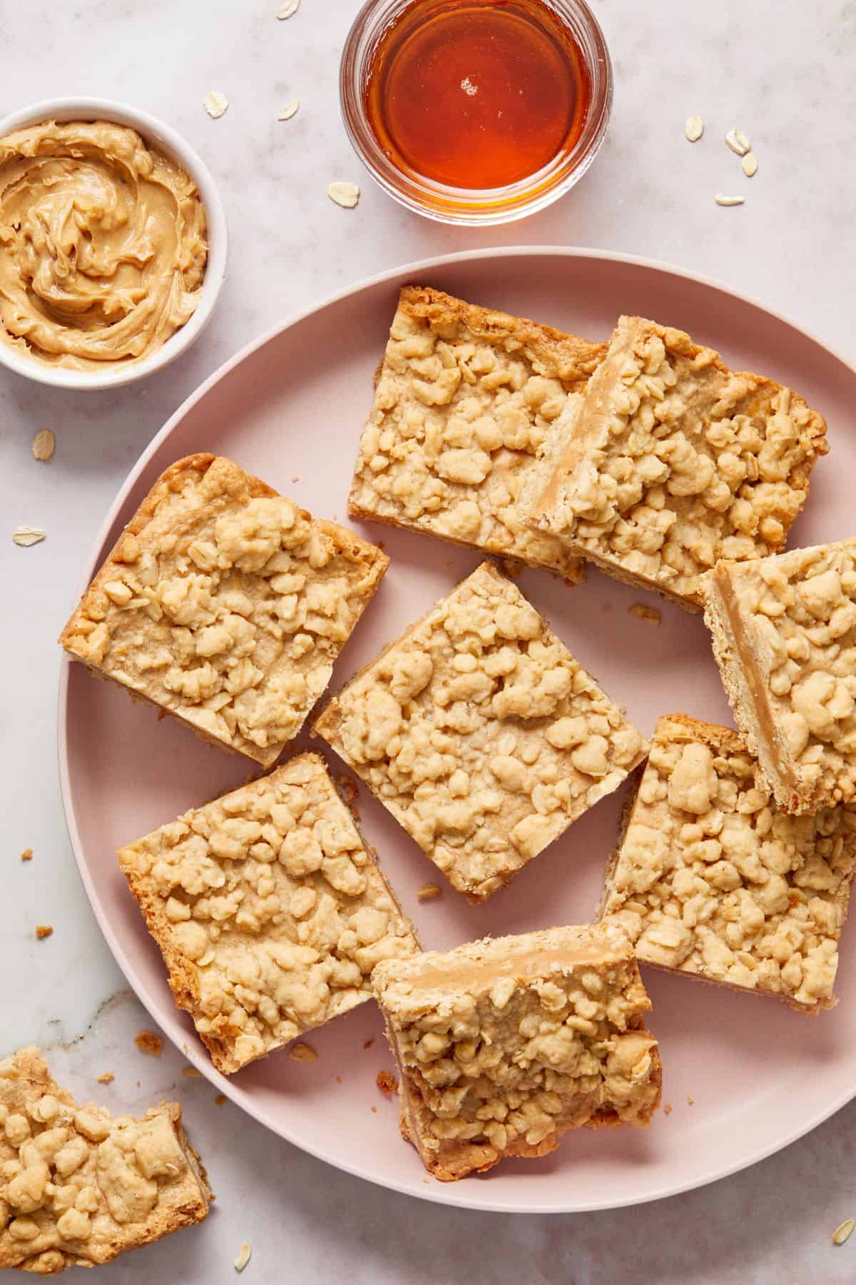 Round pink plate topped with cut squares of Peanut Butter Bars, with a small dish of peanut butter and syrup on the edge.