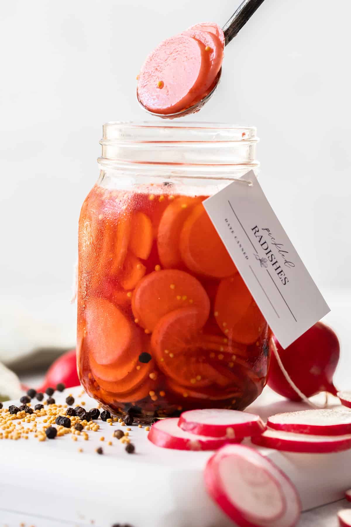 Glass jar of Pickled Radishes, with a label around the neck of the jar and a spoon lifting out some radish slices.
