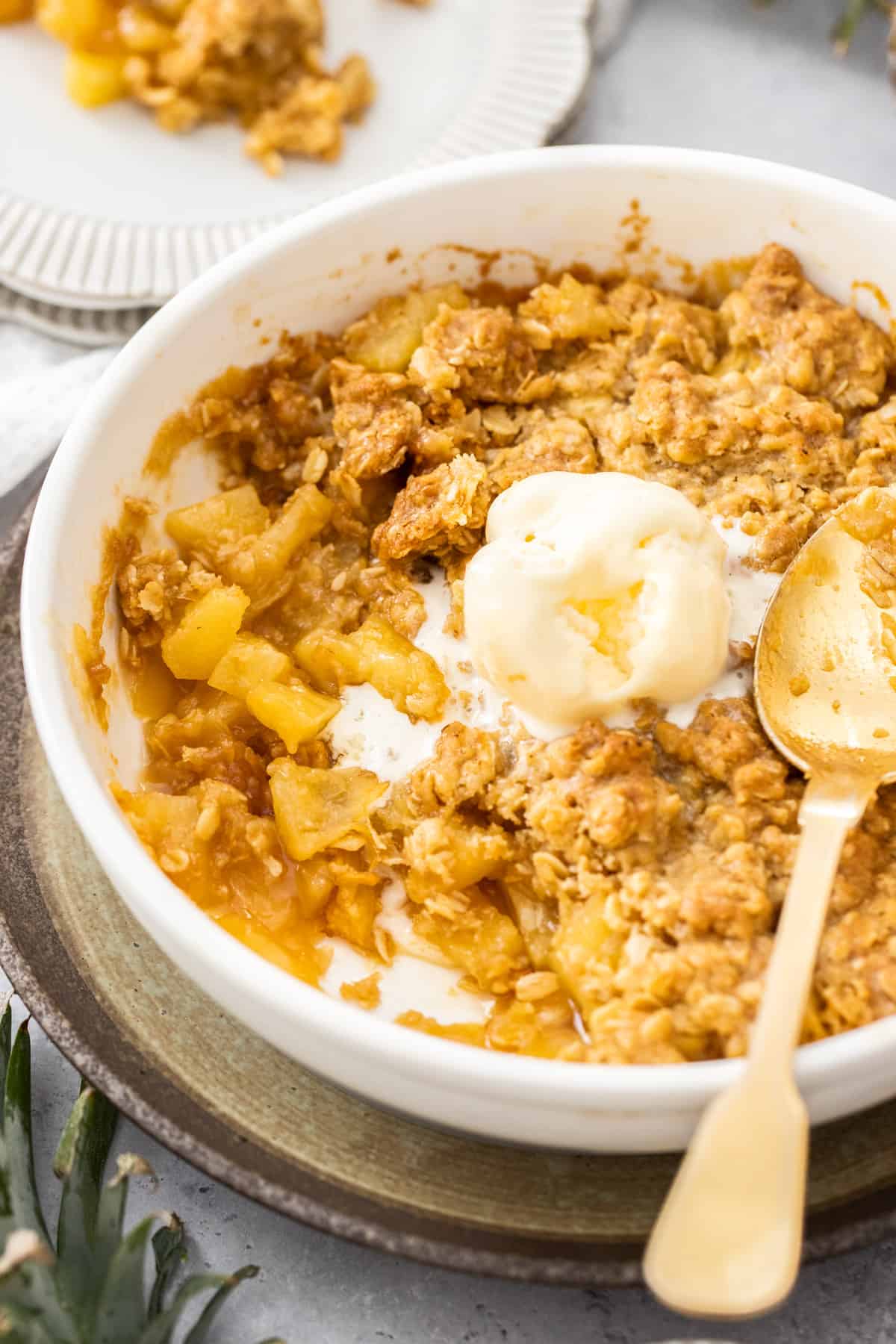 Round white dish of Pineapple Crisp, with a serve missing, a scoop of ice cream in the centre, and a gold spoon in the dish.