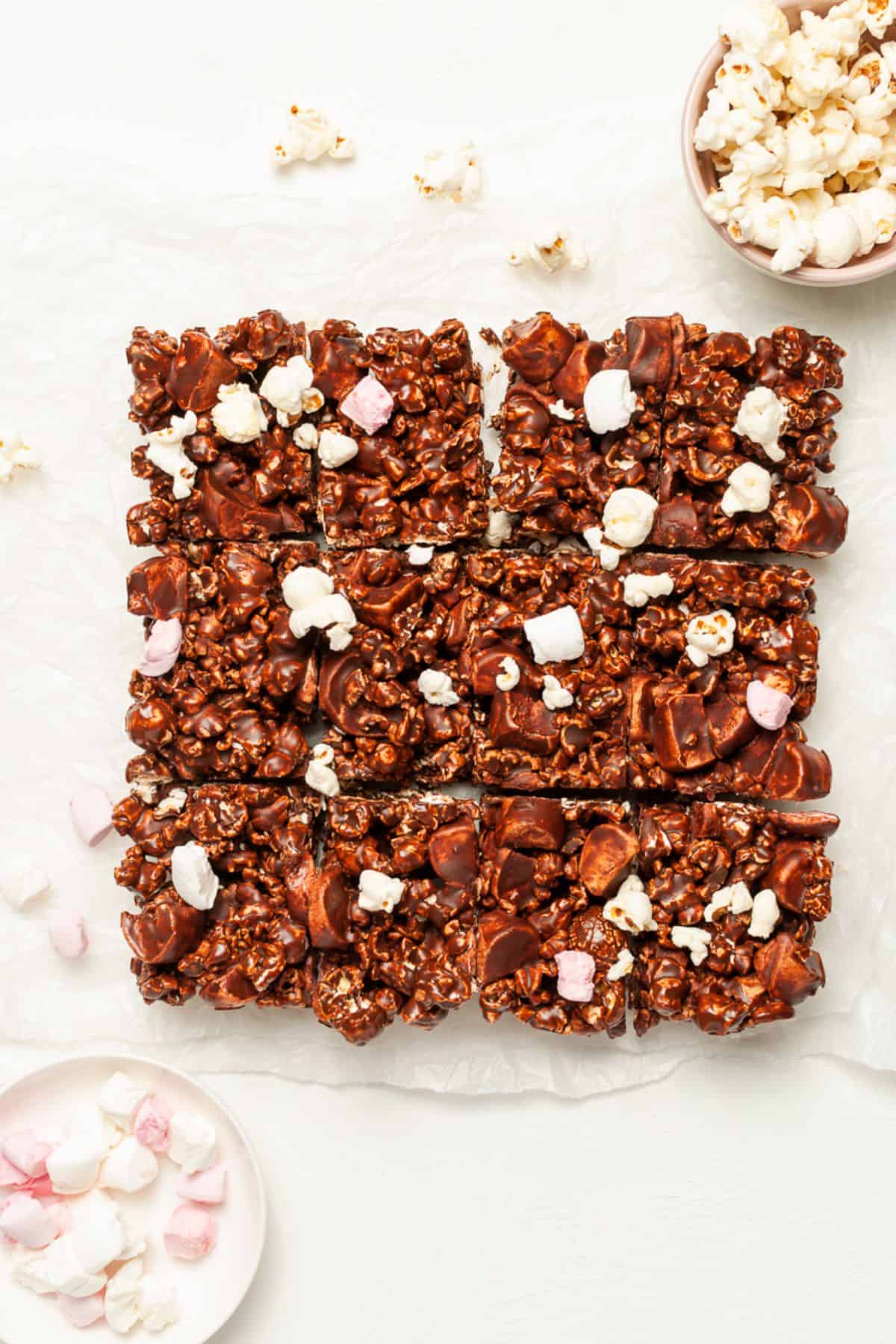 Twelve cut squares of chocolate popcorn bars on some baking paper, with some popcorn scattered around the edges.