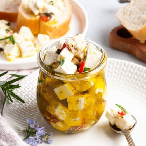 Jar of Marinated Feta, sitting on a white plate, with some slices of bread on edge.