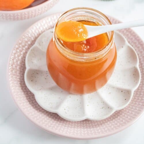Jar of Persimmon Jam, with a white spoon with jam on it resting on jar, sitting on a white plate and pink plate.