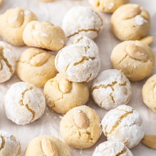 A batch of Amaretti Cookies on some baking paper - some with icing sugar and some with an almond on top.