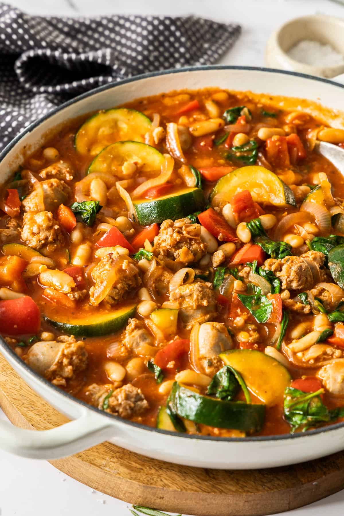 Large saucepan of Sausage Casserole with a spoon in the pan, sitting on a round wooden board.